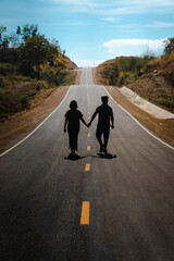 Fototapeta premium Men and women walk hand in hand on asphalt road in a rural area on a sunny day and clear blue sky clouds. There are a white border and a yellow central line. The roadside is brown and green trees.