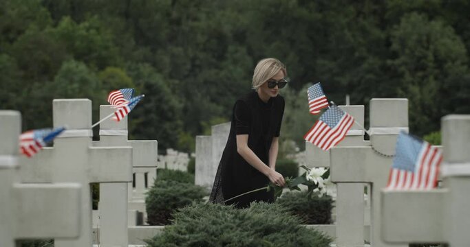 Woman putting white lily flower on grave at cemetery. Widow in dark glasses looking to camera while standing in row of stone crosses with american flags. Concept of memorial day
