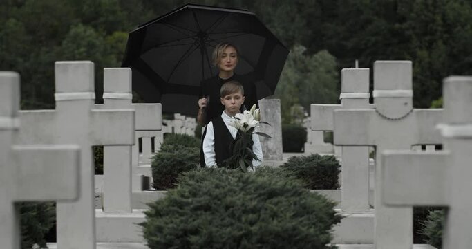 Mother in dark clothes embracing her young kid with white lily flower at cemetery.Teen boy and sad widow standing in row of stone crosses under umbrella. Concept of memorial day