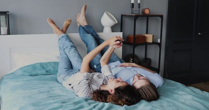 Loving smiling family couple lying on comfortable bed, making video call by mobile application, watching funny videos in social networks or taking selfie photo on smartphone in modern bedroom.