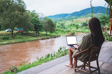 Mockup image of a young asian woman working on laptop computer while sitting by the river with mountains and nature background