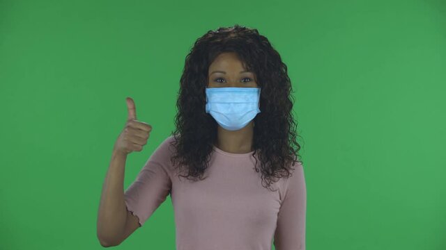 Portrait of beautiful african american young woman in medical protective face mask looking at camera and showing thumbs up gesture with both hands. Burning brunette with wavy hair in jeans and a beige