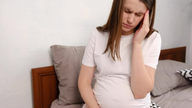 Tired young pregnant woman suffering migraine sitting on bed at home. Unhappy sad future mom holding painful head while having headache and frowning, sickness brunette female touching big belly