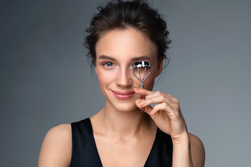 Beautiful woman using eyelash curler on long eyelashes. Photo of woman with perfect makeup on gray background. Beauty concept
