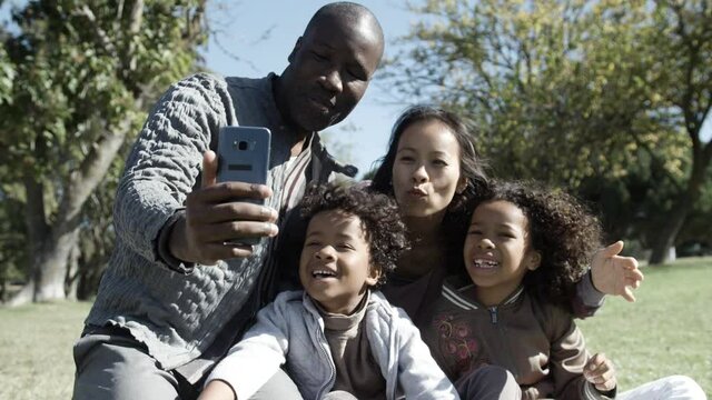 Happy Mixed-race Family Sitting On Lawn And Having Video Chat Via Smartphone. African American Dad Holding Phone, Asian Mom And Kids Smiling, Kissing Air And Waving. Family And Holiday Concept