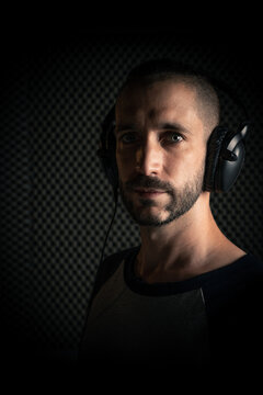 Hombre con peinado moderno y ojos azules, y camiseta gris de mangas azul marino con auriculares en un estudio de grabación de sonido mirando a cámara con luz de flash
