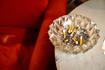 Cenicero lleno de colillas y ceniza junto a un sillón rojo y mesa de madera sucios