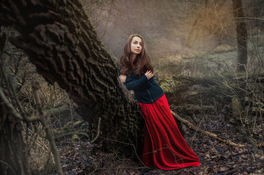 Pensive, dreamy beautiful young woman in the autumn forest. Autumn nature. A beautiful girl in a red dress on the shore of an autumn reservoir. Young woman in sexy red dress in the forest
