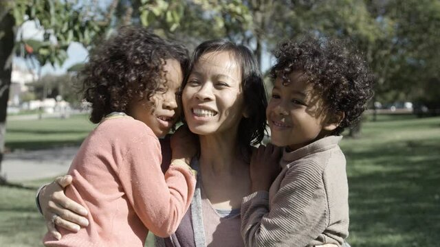 Happy Asian mom talking, hugging and kissing kids in city park. Little cute mixed-race son and daughter embracing mother and smiling. Front view. Static camera. Family, love and weekend concept