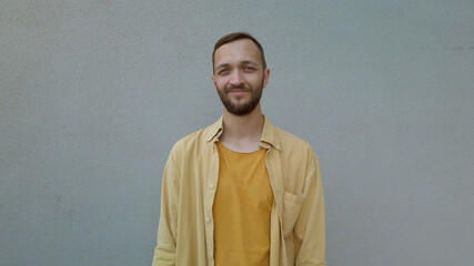 Front view portrait of handsome bearded man smiling and standing against a grey wall. Male expressing emotion