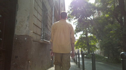 Fototapeta na wymiar Back view of adult male traveler in yellow shirt walks along a narrow old street. Tourism during quarantine