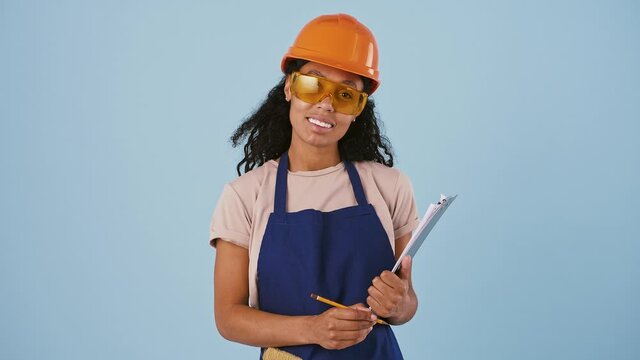 Dark-skinned girl in hard hat and protective goggles. She nodding her head and smiling, holding clipboard and pencil. Posing on blue background