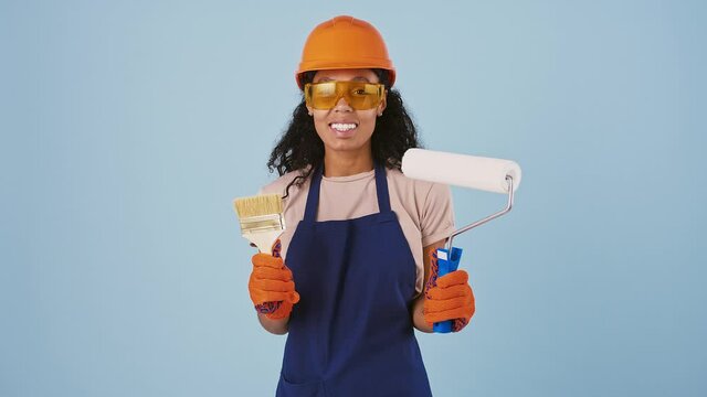 Afro-american lady worker in safety clothes, hard hat, protective goggles, gloves. Smiling, holding paint roller and brush. Posing on blue background