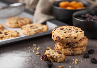Homemade organic oatmeal cookies with raisins and apricots with baking tray on grey wooden background. Black bowl of raisins and dried apricots