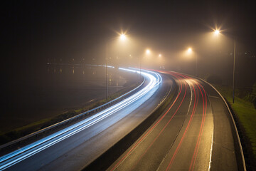 Car light trails on a highway at night, rounding a bend into the mist