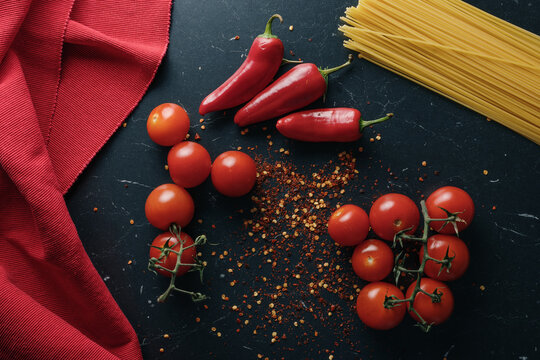 Dark kitchen countertop with a kitchen towel in red cotton and cherry tomatoes, tuft of pasta spaghetti, ground hot peppers, spaghetti and capsicum