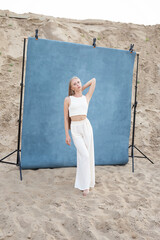 beauty portrait outdoors on sand in front of blue background, pretty female posing. attractive caucasian girl with long hair looking at camera. beautiful woman in white clothes, elegant fashion style