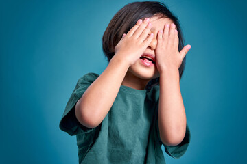 Happy asian little kid blocking face with both hand, empty space in studio shot isolated on colorful blue background. Education concept for school.