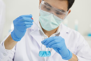 Male Asian Scientist wears Mask working in Lab while Droping Solution  to Erlenmeyer Flask. Blue...