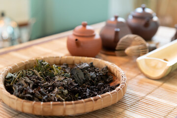 Tea leaves placed in a basket and 3 small ceramic kettles for brewing light brown and dark brown teas, arranged on a Japanese bamboo mat in a coffee shop.