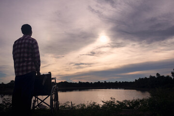 Naklejka na ściany i meble Silhouette young disabled man seeing river background.He is raise a hand to and sitting on wheelchair.despair,lonely,hope.Photo concept depression and Patient.