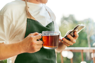 Crop close-up man drinks hot tea and holds a smartphone in his hands on a sunny day. Business success. Stay at home concept.