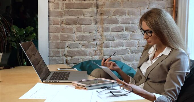 The charming young blonde businesswoman in glasses is working with documents while sitting at the laptop in the office.