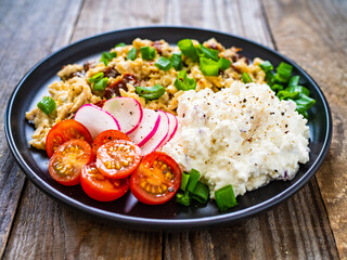 Delicious breakfast - scrambled eggs with fried sausages, cottage cheese and vegetables served on black plate on wooden table
