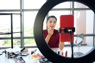 Beautiful Asian woman blogger hand holding lipstic and recording on red smartphone for make up tutorial video on website. Marketing in social media and Work from Home concept.