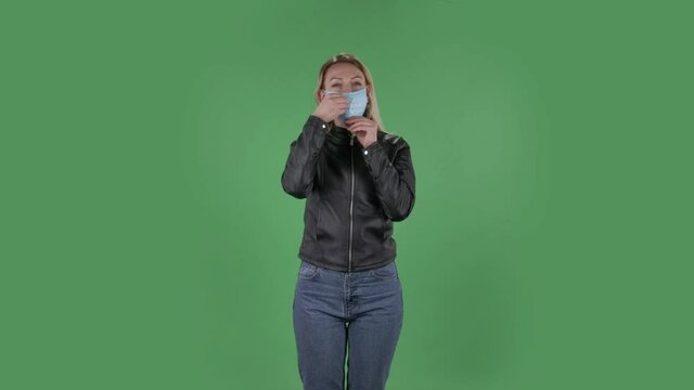 Portrait of beautiful young woman puts on medical mask. Blonde with loose hair in a black jacket and jeans on a green screen in the studio. Health Protection Corona Virus Concept.