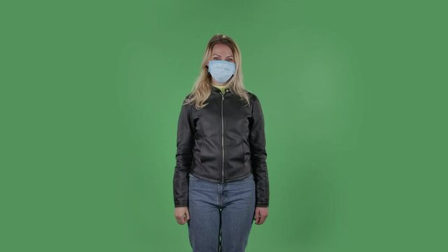 Portrait of beautiful young woman in medical mask is looking at camera and joyfully making sign ok. Blonde with loose hair in a black jacket and jeans on a green screen in the studio. Health