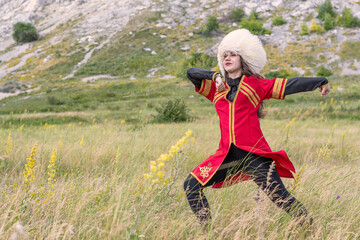 A young cheerful girl in a national costume is dancing a fiery dance on a green field at the foot of the mountain
