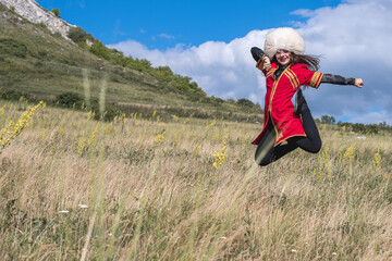 A cheerful young girl in a red suit soars over a green field, dancing a national dance against a blue sky