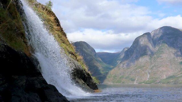 Waterfall falling into norwegian fjord Aurlandsfjord. Close bright white water view on wild mountains nature of Norway. Sunny travel day.