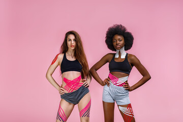 Kinesiology taping. Two young female athletes on pink background with kinesiology tape on neck,shoulder, khee, tummy and hips. Fat lose, cellulite removal, sport physical therapy,recovery concept.