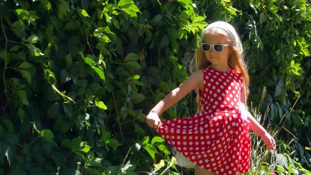 Beautiful happy girl in a red polka dot dress smiling on wild grape background. Cute joyful child with long blonde hair in sunglasses and white hat on green natural backdrop outdoor.