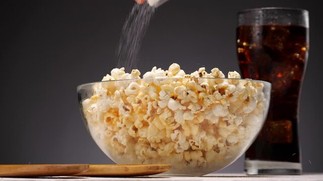 Unrecognizable person spilling salt on popcorn then mixing snack in bowl near glass of soda on gray background