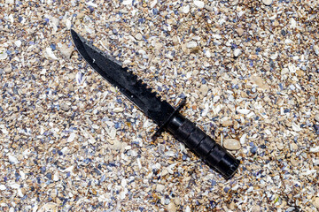 black camping knife on the beach with colorful sea stones and clear sea. camping and holiday concept background