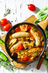 Stew of beans, vegetables and smoked sausages with tomato sauce in the pan on wooden table. Top view flat lay background. Copy space.