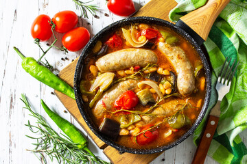 Stew of beans, vegetables and smoked sausages with tomato sauce in the pan on wooden table. Top view flat lay background.