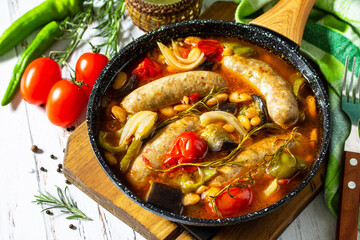 Stew of beans, vegetables and smoked sausages with tomato sauce in the pan on wooden table.