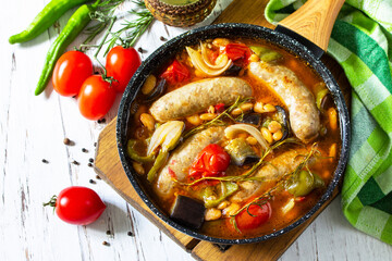Stew of beans, vegetables and smoked sausages with tomato sauce in the pan on wooden table.