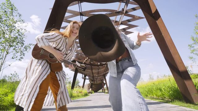 attractive cheerful women with ukulele guitar and cowboy hat dance and sing under openwork canopy in contemporary decorated city garden on sunny day