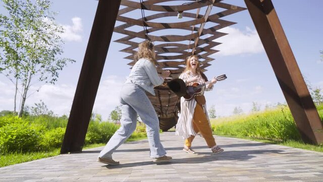 active long haired girls with ukulele guitar and cowboy hat dance and sing under openwork canopy having fun in decorated city park in sunny morning