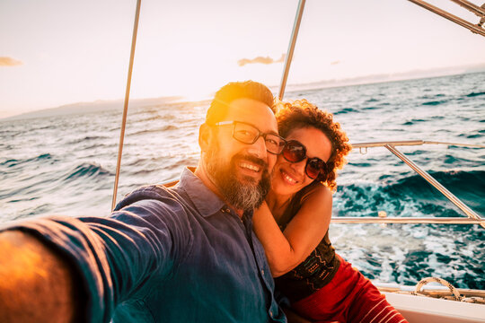 Adult happy caucasian couple take selfie picture yachting on a boat enjoying the sunset on the ocean - sun and sky in background - joyful and freedom people in love concept