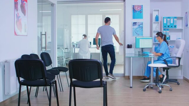 Nurse with face mask for covid-19 in hospital waiting area checking senior man temperature with temperature scanner. Assistant helping invalid senior woman in wheelchair.
