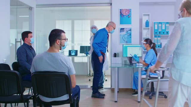 Medic with visor against covid 19 calling patient in consultation room. Senior man with face mask in hospital waiting room talking with nurse. Disabled woman with zimmer frame.