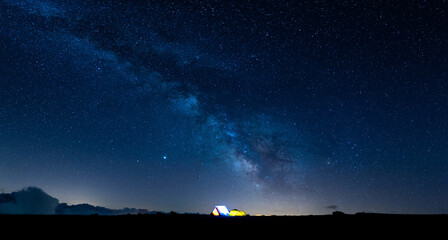 Camping in the mountains under the starry sky and the vastness of the universe. Panoramic landscape of the Milky Way in the starry night sky with small bright tents below.