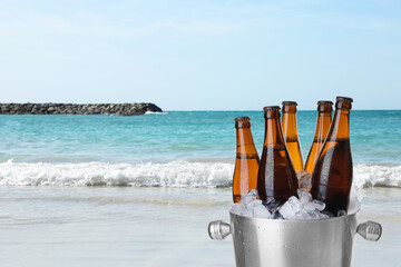 Bottles of beer with ice cubes in metal bucket against ocean and sandy beach. Space for text