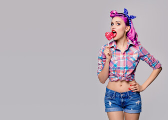 Portrait image of woman licking heart shape lollipop dressed in pinup style. Purple girl in retro fashion and vintage concept. Grey color background. Copy space for some text.